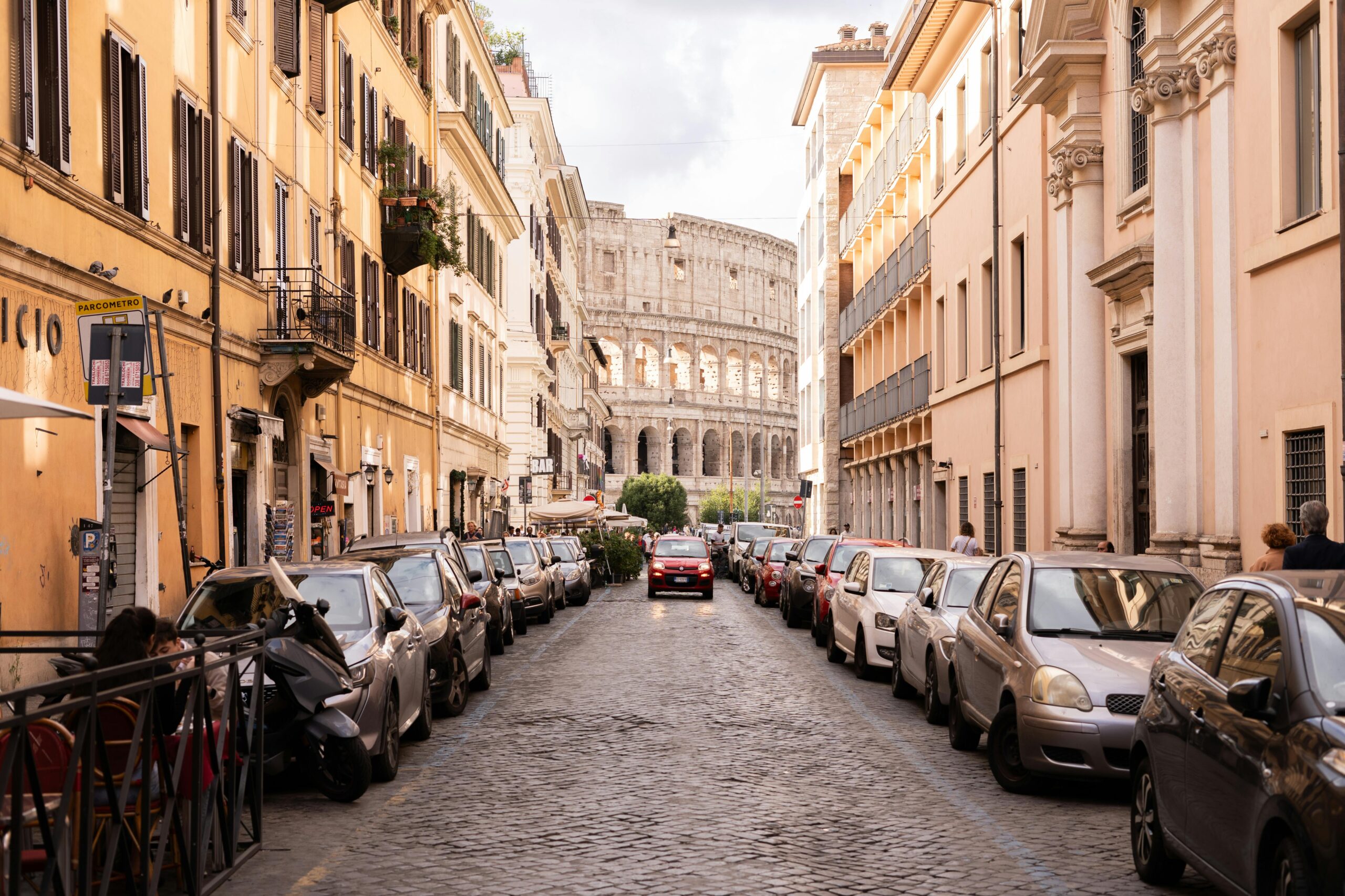 Street view in Rome, featuring cars lined up and the iconic Colosseum in the background.